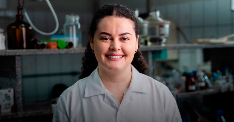 Raquel Moreira é uma pesquisadora de cabelos cacheados e escuros, vestindo um jaleco branco. Atrás dela, aparecem vidraçarias de laboratório