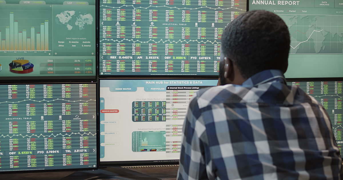 Imagem é uma fotografia que mostra um homem de costas analisando dados do mercado financeiro expostos em diversos monitores à sua frente