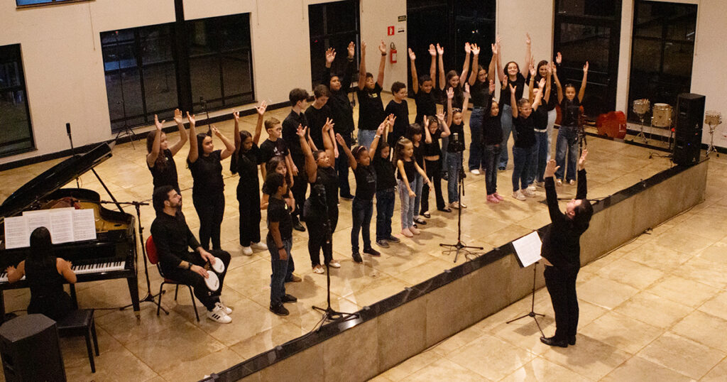 Grupo de crianças num palco vestidos de preto e com as mãos para cima. Na frente delas o maestro e ao lado uma pessoa sentada em frente a um piano.