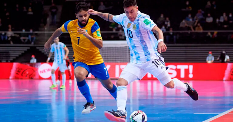 Dois homens, um branco com uniforme branco e azul e um negro com uniforme azul e amarelo, brigando pela bola em uma partida de futsal em um ginásio