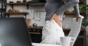 Mulher levantando da cadeira de mesa de cozinha com a mão encostada na lombar