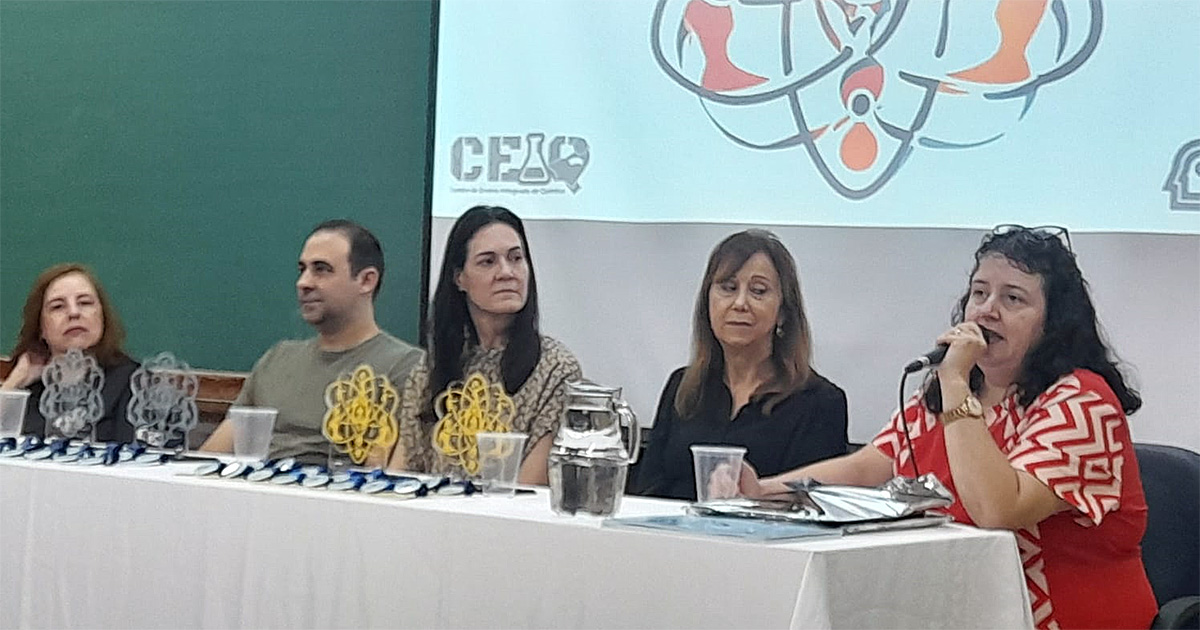 Mesa coberta por uma toalha branca, com troféus e medalhas em cima. Atrás da mesa, quatro mulheres e um homem sentam-se à frente de um telão.