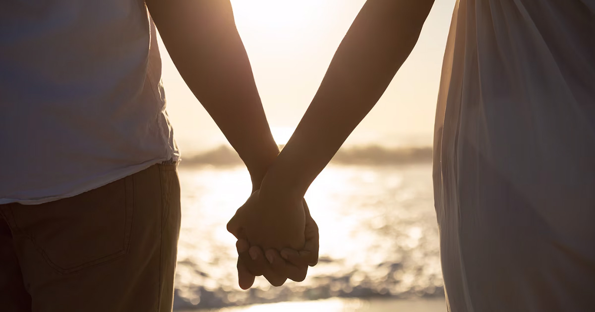 Imagem é uma fotografia de um casal de mãos dadas em frente à praia, com destaque para as mãos do casal.