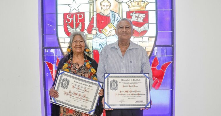 Mulher indígena ao lado de homem negro, ambos posando para foto segurando diplomas da USP, na frente de um grande vitral com o brasão da USP