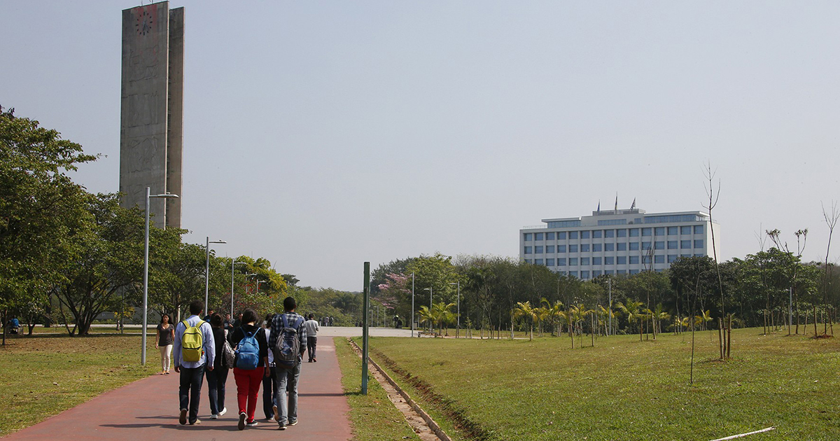 Estudantes caminhando por praça cercada de grama. À direita, está a torre do relógio e, à direita, um prédio baixo.