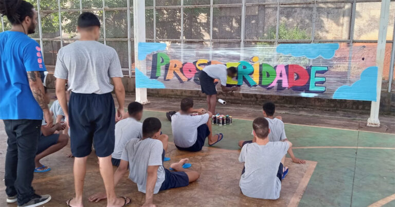 Imagem com cinco meninos sentados no chão, um em pé e outro agachado na frente de todos e de costas, grafitando a palavra prosperidade toda colorida. todos usam camiseta cinza e calção azul escuro