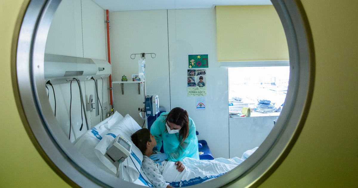 Na foto, uma enfermeira atende uma criança deitada em cama hospitalar. A imagem foi feita através de uma janela redonda.