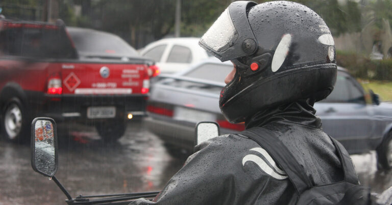 A imagem mostra detalhe de um motociclista parado no trânsito em um dia chuvoso. Ele está usando um capacete e uma capa de chuva preta. Ao fundo, há um veículo vermelho e outro cinza