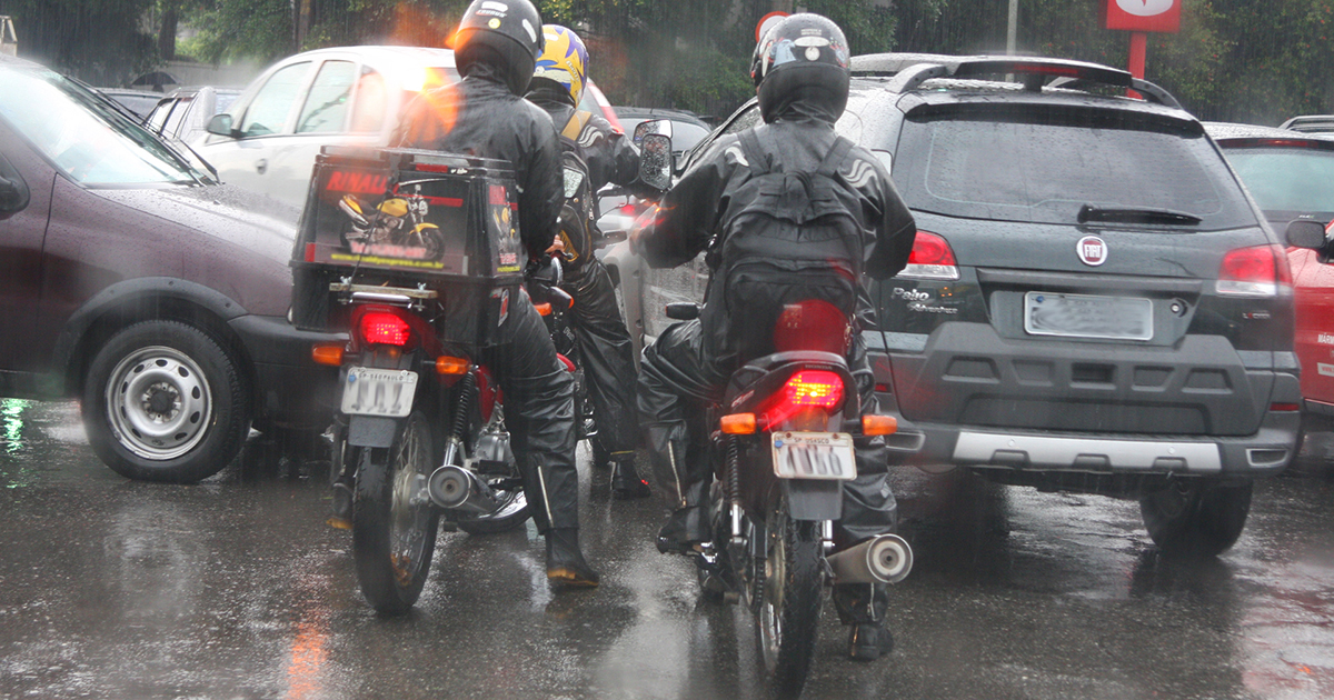 A imagem mostra dois motociclistas em um dia chuvoso, tentando driblar o trânsito. Eles estão usando capas de chuva e estão parados entre carros no trânsito