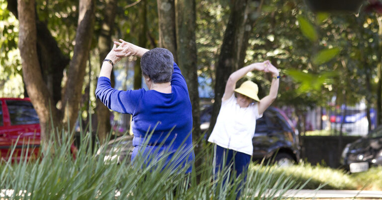 Duas mulheres brancas idosas com um fundo de uma rua bem arborizada com três carros estacionados