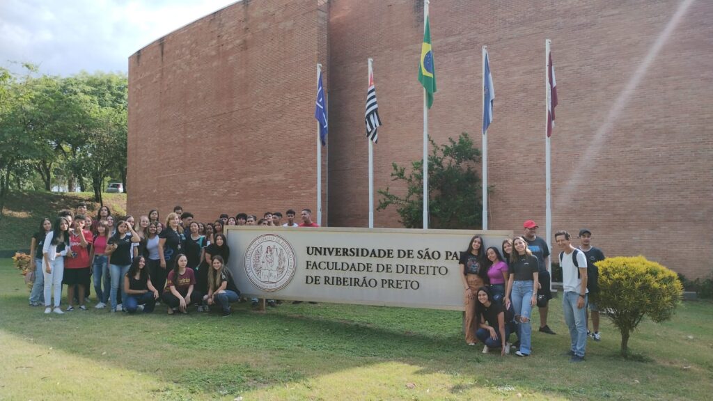 A imagem mostra um grupo grande de estudantes reunidos em frente a um edifício de tijolos aparentes, Na parte central, há uma placa grande com o nome da instituição, “Universidade de São Paulo - Faculdade de Direito de Ribeirão Preto”, e o símbolo oficial do lado esquerdo. Várias bandeiras estão posicionadas em mastros ao fundo, incluindo as bandeiras do Brasil, do estado de São Paulo e outras. O grupo é formado por jovens, muitos deles sorrindo e fazendo poses casuais, alguns agachados e outros em pé. O local tem uma área verde com grama e árvores ao redor, e o céu está parcialmente nublado, mas ensolarado.