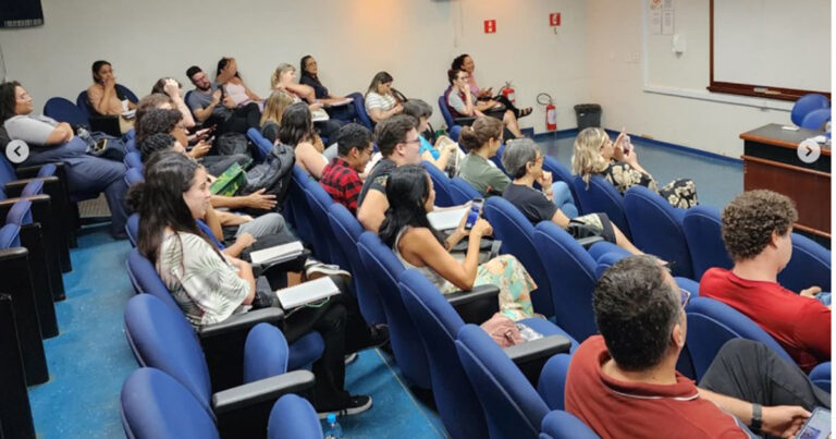 A imagem mostra um auditório com cadeiras azuis ocupadas por várias pessoas. Elas estão sentadas, algumas anotando em cadernos, outras usando celulares ou apenas prestando atenção na apresentação de um evento acadêmico. O espaço é iluminado e tem paredes brancas, além de equipamentos de segurança como extintores de incêndio visíveis. Um púlpito ou mesa para palestrante aparece no canto direito.