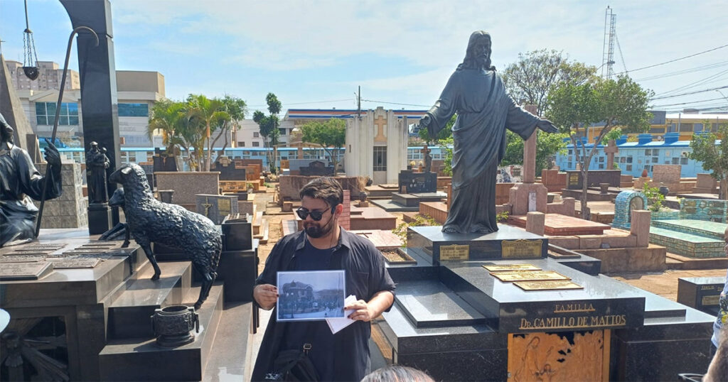 Imagem dentro de um cemitério, cercado com um muro azul, com túmulos ao redor de um homem branco com camiseta escura e óculos escuros. do lado direito uma estátua alta de um santo com os braços abertos e do lado esquerdo uma estátua de um cordeiro