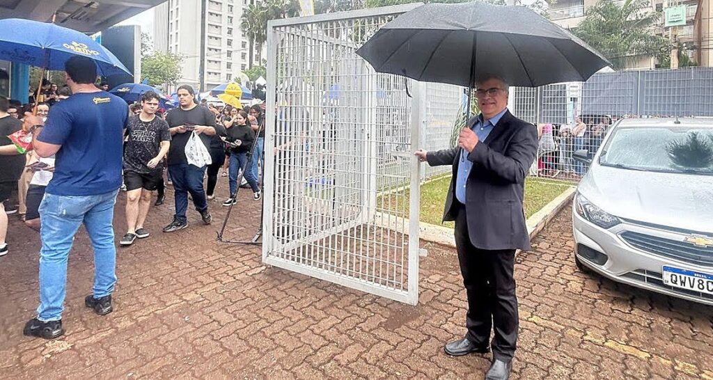 Homem branco, segurando um guarda-chuva e abrindo um portão branco.