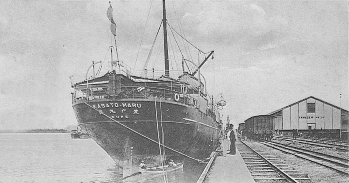 Foto em preto e branco mostra a proa de um navio ao lado de uma linha férrea. À direita, uma construção de madeira e pessoas circulando; à esquerda, um braço d'água natural com um barco ao fundo