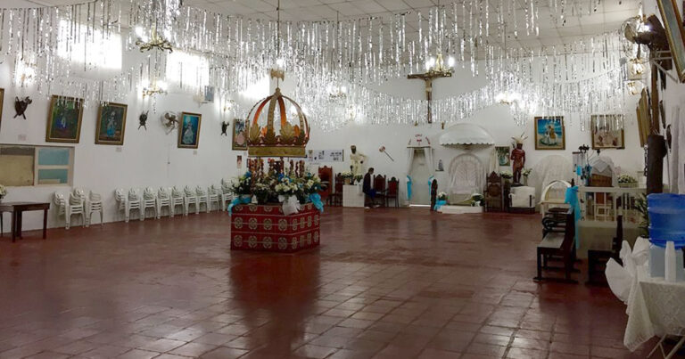 Salão do Axé Ilê Obá é decorado com arranjos de flores, estátuas de caboclos, quadros de orixás e um crucifixo cristão. Ao centro, um altar coberto por flores, laços e uma grande coroa.