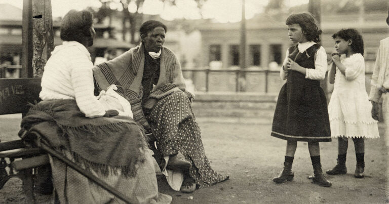 Sentadas em um banco público, duas mulheres negras estão conversando. Em pé, duas crianças brancas as observam atentamente. A foto é antiga, datada de cerca de 1910.