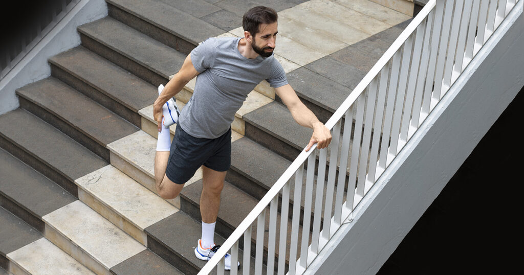 A imagem apresenta um homem com roupas esportivas fazendo alongamento da perna direita numa escada, se apoiando no corrimão.