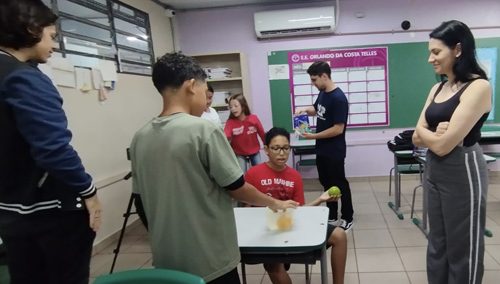 No centro, três crianças fazem um experimento com duas bolas e um recipiente com água. À direita, uma aluna da USP observa as crianças, sorrindo. Ao fundo, outro universitário faz outro experimento com outras crianças.