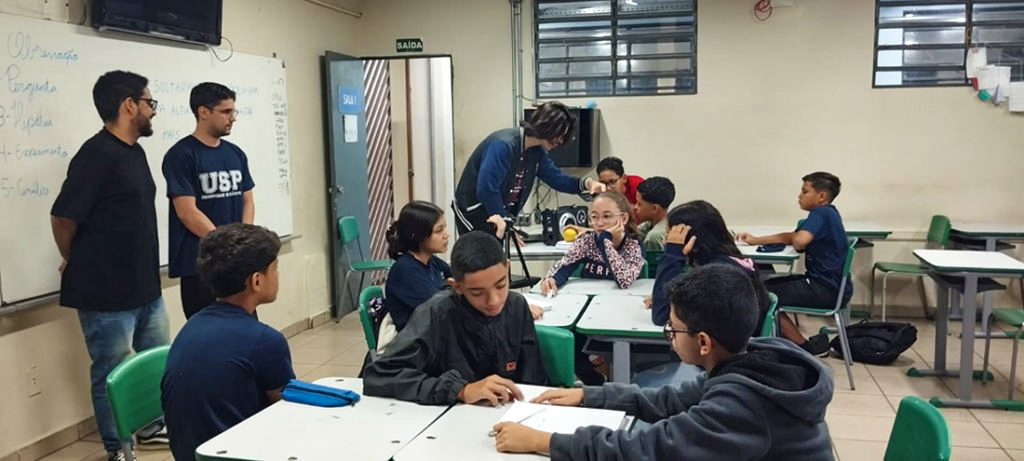 Em uma sala de aula, oito crianças estão divididas em grupos e conversam entre si. À esquerda, observando os alunos, estão os organizadores do projeto. Um dos universitários conversa com um dos alunos ao fundo.