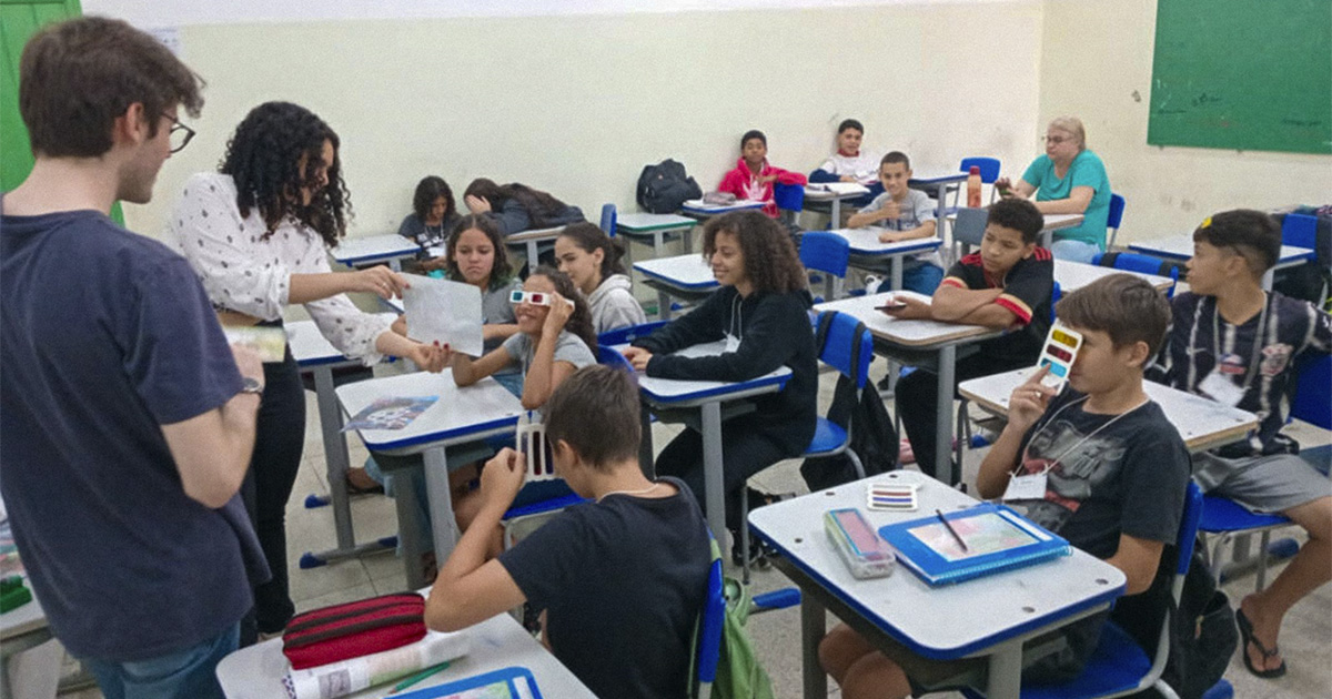 Em uma sala de aula, várias crianças estão sentadas, olhando em direção a uma menina, que usa um óculos 3D enquanto vê uma imagem colorida e sorri. À esquerda, estudantes da USP mostram papéis com imagens coloridas.