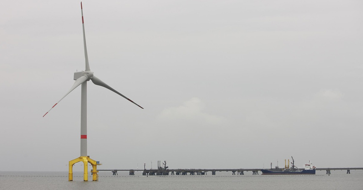 Imagem é uma fotografia de uma torre de energia eólica no mar com um braço de atracação de navios.