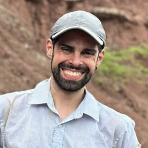 Homem branco adulto com barba e cabelos escuros e curtos, que estão cobertos por um boné. Está em um ambiente de terra, vestindo uma camisa azul clara e está sorrindo.