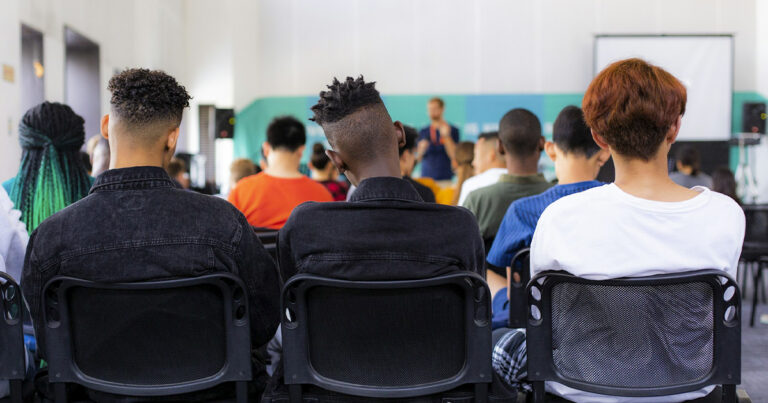 Jovens pretos, pardos e brancos assistem a uma palestra, sentados em uma sala de aula