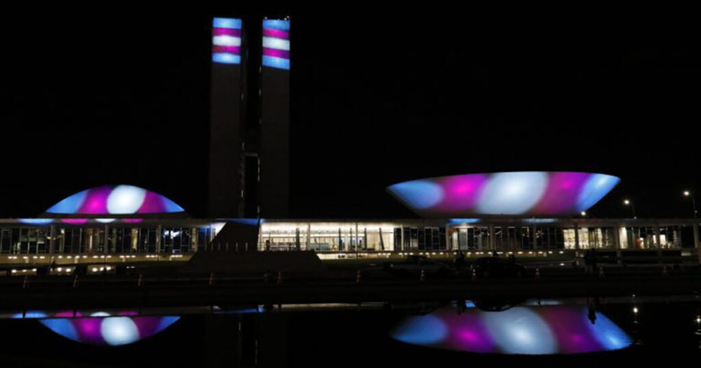 A imagem mostra o Congresso Nacional iluminado com as cores rosa, azul e branco