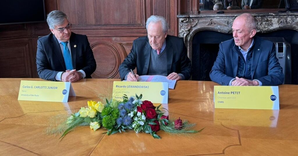 A imagem mostra três homens sentados à mesa em cerimônia de assinatura. à frente do homem do meio há um buquê de flores