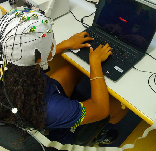 Mulher fotografada de cima usando uma touca branca com eletrodos e fios. Ela usa um uniforme azul e está sentada em frente a um notebook preto com a palavra "vermelho" escrita na tela