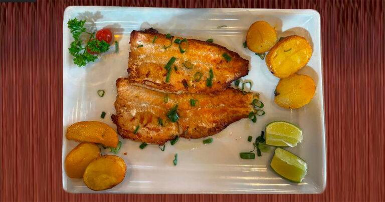 travessa branca com dois filés de peixe (cor amarela/dourada), acompanhado de batatas assadas, limão, tomate cereja e um galhinho de salsinha verde.