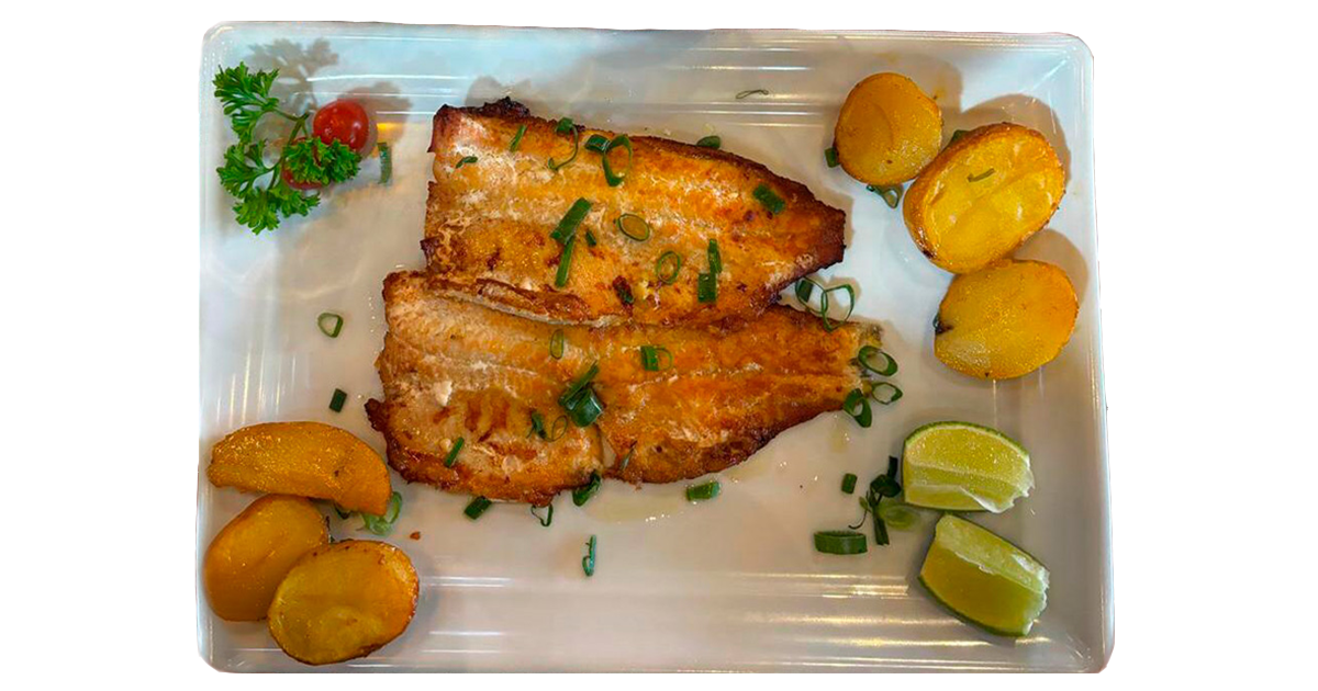 travessa branca com dois filés de peixe (cor marela/dourada), acompanhado de batatas assadas, limão, tomate cereja e um galhinho de salsinha verde.