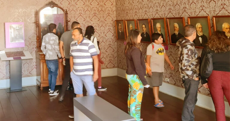 Oito pessoas numa sala de museu olhando quadros de personalidades e móveis antigos