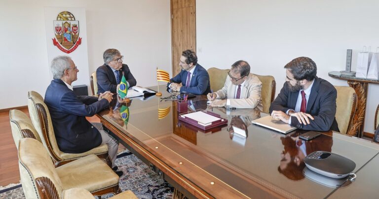 Homens sentados ao redor de uma mesa de reuniões. Na mesa, pequenas bandeiras do Brasil e Catalunha. Ao fundo, totem com o brasão da USP