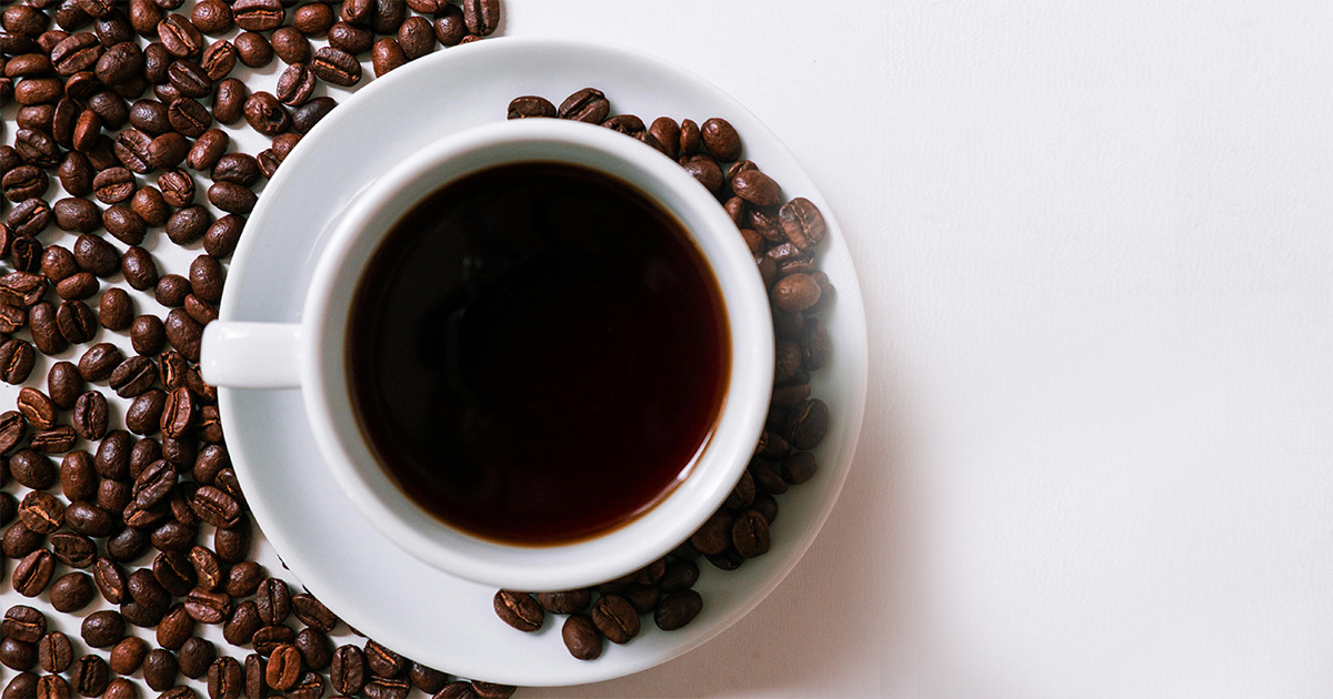 Imagem vista do alto de uma xícara de café cheia até a borda com a bebida e, espalhados em volta, estão grãos de café