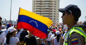 manifestação com centenas de pessoas na rua; em destaque, uma pessoa usando boné e camiseta brancos segura uma bandeira da Venezuela do lado esquerdo da imagem, e uma autoridade da polícia nacional bolivariana de perfil do lado direito da imagem