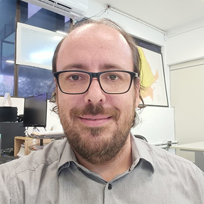 Homem branco, ainda jovem, barba e bigode, cabelo escasso na testa e usando óculos. Vete camisa bege.