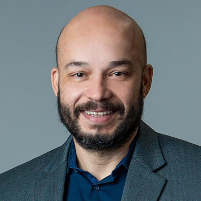 Homem branco, calvo, de barba, vestido de paletó e camisa escuros, esboçando um leve sorriso.