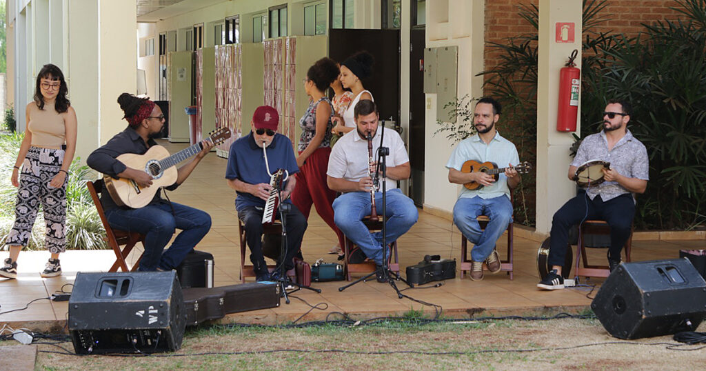 A imagem mostra um grupo de cinco músicos tocando ao ar livre em um espaço coberto. Os músicos estão sentados em cadeiras, tocando diferentes instrumentos: violão, melódica, clarinete, cavaquinho e pandeiro. Há caixas de som no chão à frente deles. Atrás do grupo, algumas pessoas caminharam e observaram a apresentação. O ambiente tem um piso de cerâmica e uma parede com plantas ao fundo, além de um extintor de incêndio fixado na parede. 