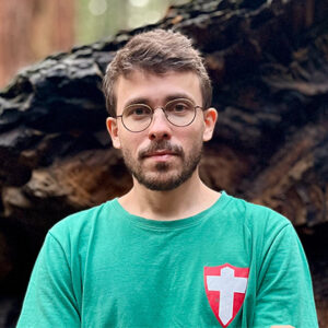 Homem branco de barba e cabelos claros, usando óculos e camiseta verde, com uma cruz estampada na camiseta.