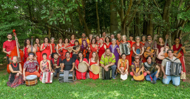 Músicos de uma orquestra com seus instrumentos posando para a foto, tendo árvores ao fundo.