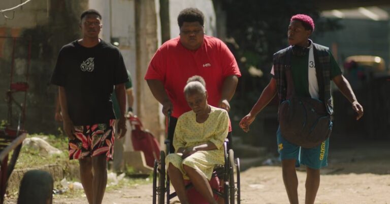 3 jovens andam por uma rua de chão batido, um deles empurra uma senhora em uma cadeira de rodas