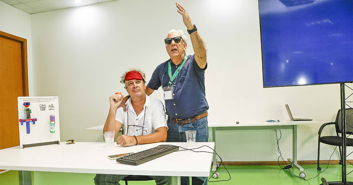 dois homens atrás de uma mesa. Um sentado de e o outro de pé. A mese está com um teclado e alguns protóticos de equipamentos para auxiliar as pessoas com baixa visão.