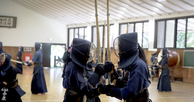 A imagem mostra uma aula de kendo, uma arte marcial japonesa que utiliza espadas de bambu chamadas "shinai". Dois praticantes estão no centro, vestindo armaduras tradicionais chamadas "bogu", que incluem capacetes com grades metálicas para proteção do rosto. Eles estão em uma posição de combate, segurando suas espadas e olhando um para o outro. No fundo, há outros praticantes treinando em um dojo espaçoso, com janelas grandes que permitem a entrada de luz natural. O ambiente tem um piso de madeira, e há uma atmosfera disciplinada e focada.