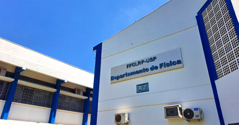 A imagem mostra a fachada de um prédio branco com detalhes em azul, identificado como o "Departamento de Física" da FFCLRP-USP (Faculdade de Filosofia, Ciências e Letras de Ribeirão Preto, da Universidade de São Paulo). Há um letreiro em relevo com o nome do departamento e a identificação "B2". O céu está azul e claro. Algumas janelas têm grades metálicas e há aparelhos de ar-condicionado instalados na parede.