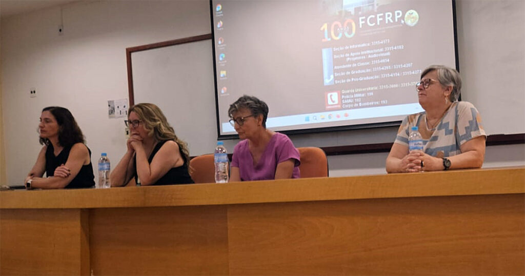 A imagem mostra quatro mulheres sentadas em uma mesa longa de madeira em uma sala de aula ou auditório e e há garrafas de água à sua frente. No fundo, há um projetor exibindo uma tela com informações sobre um evento incluindo o número "100" e a sigla "FCFRP". A sala tem paredes claras, um quadro branco.