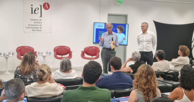 A imagem mostra um evento em um auditório com várias pessoas assistindo a uma apresentação. Dois homens estão em pé na frente da sala. Há uma tela atrás deles exibindo um slide de apresentação com uma imagem e texto. No fundo, há uma parede branca com um painel que tem o logotipo e o nome do "Instituto de Estudos Avançados" (IEA) da Universidade de São Paulo (USP). Algumas cadeiras vermelhas vazias estão posicionadas à frente. O público é composto por homens e mulheres, sentados.