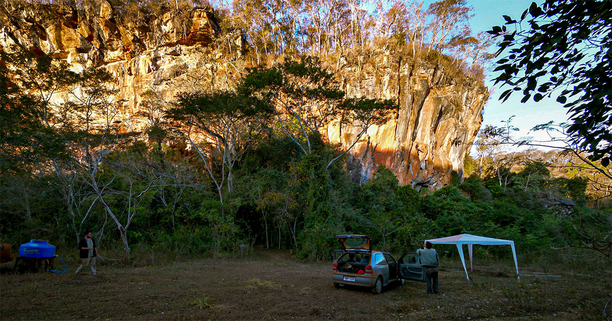 Vista de um maciço rochoso ao entardecer. Nos pés do maciço, algumas árvores; à esquerda, uma caixa d'água azul que permite o peneiramente do material escavado e à direita uma tenda branca ao lado de um veículo. Dois homens circulam pelo local