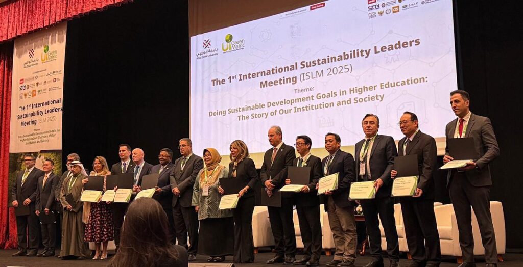 A imagem mostra um grupo de pessoas em um palco, segurando certificados e pastas, durante um evento chamado "The 1st International Sustainability Leaders Meeting (ISLM 2025)". O fundo do palco exibe um grande banner com o nome do evento. Os participantes estão vestidos formalmente, alguns com ternos e gravatas, enquanto outros usam vestimentas tradicionais.